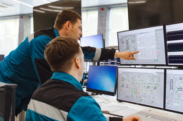 Dois profissionais de uniforme azul numa sala de controle, um deles apontando para monitores com telas de supervisão da operação